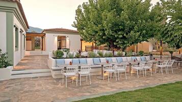 Un patio al aire libre con sillas y mesas blancas, suelo de piedra y un gran árbol que da sombra.