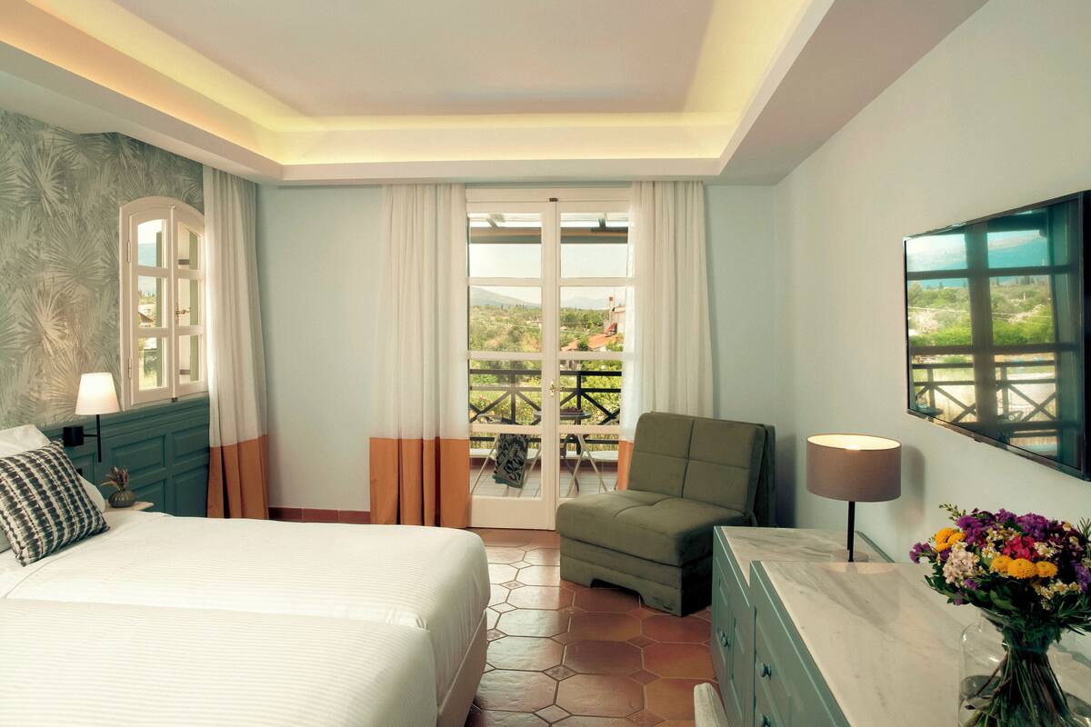 Een hotelkamer met een bed, een groene fauteuil, een bureau met lamp, een bloemenvaas en uitzicht op het groen door een schuifdeur.