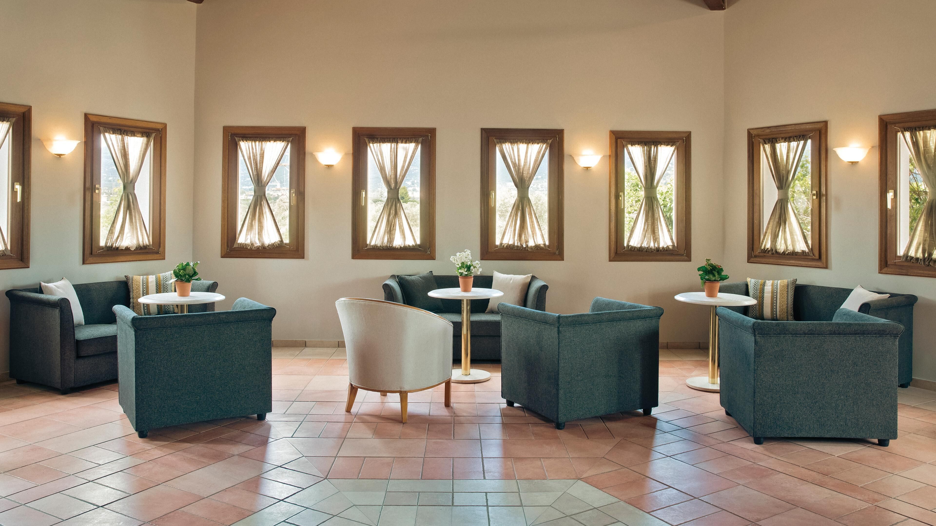 Una habitación amplia con varias zonas de estar, incluyendo sofás y sillones, dispuestos alrededor de una mesa de centro. Hay seis ventanas con cortinas, que dejan entrar luz natural.