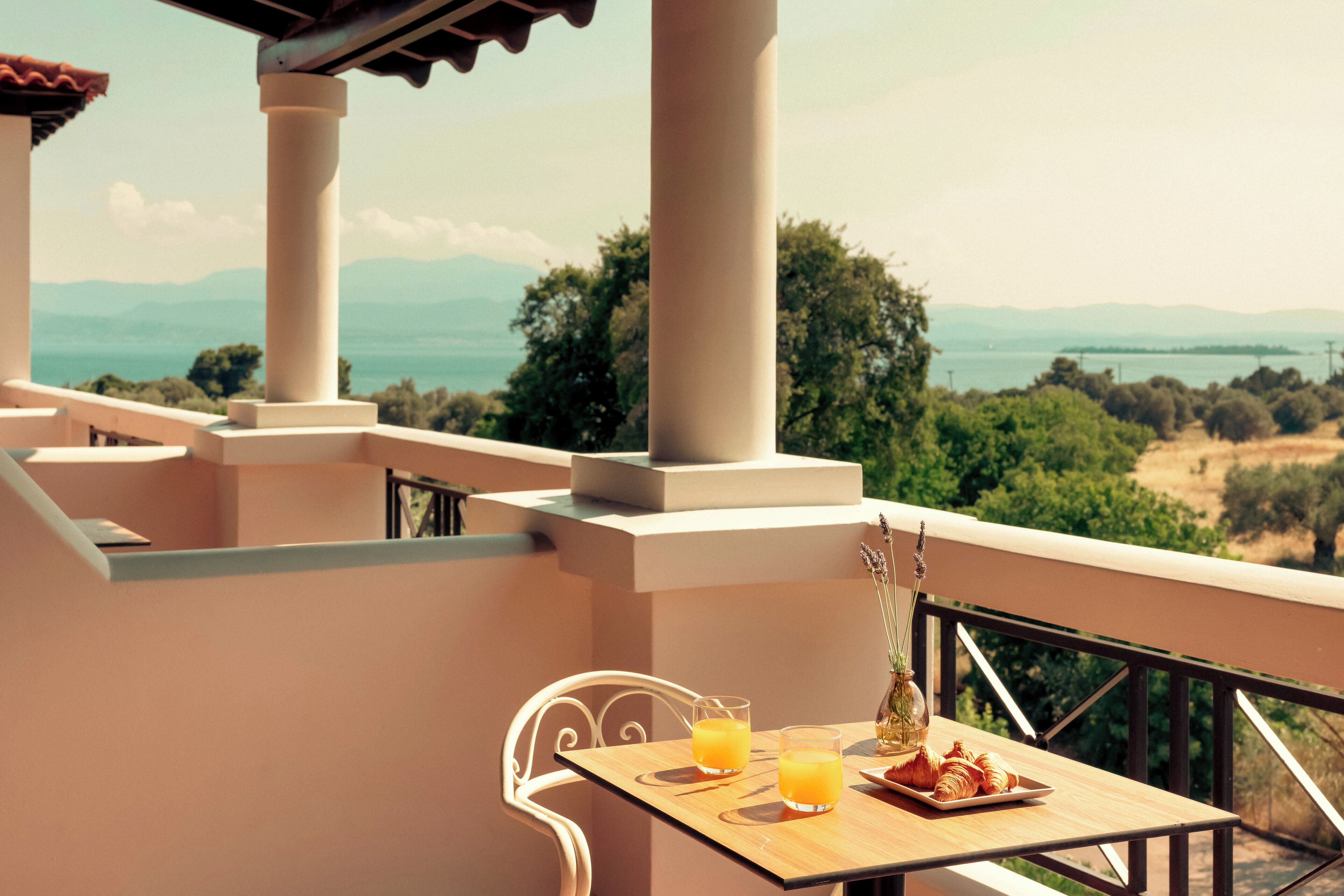 A balcony with a table set for two, two glasses of orange juice, a plate of croissants, and a vase with flowers.