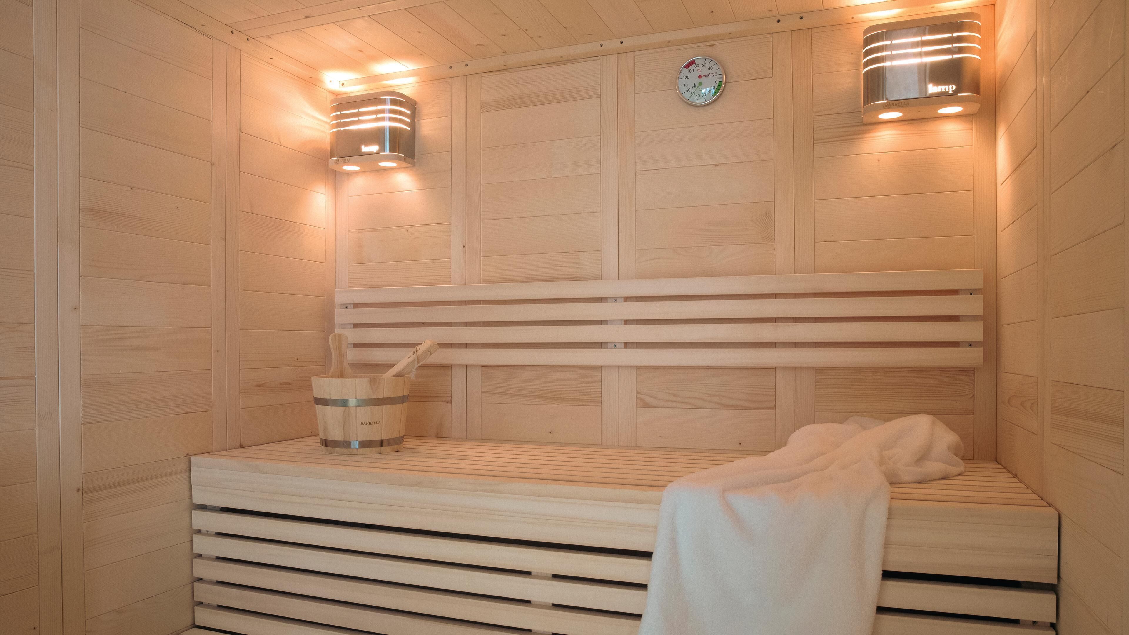 A sauna with wooden walls, a bench, a bucket with a mallet, and two wall-mounted lights.