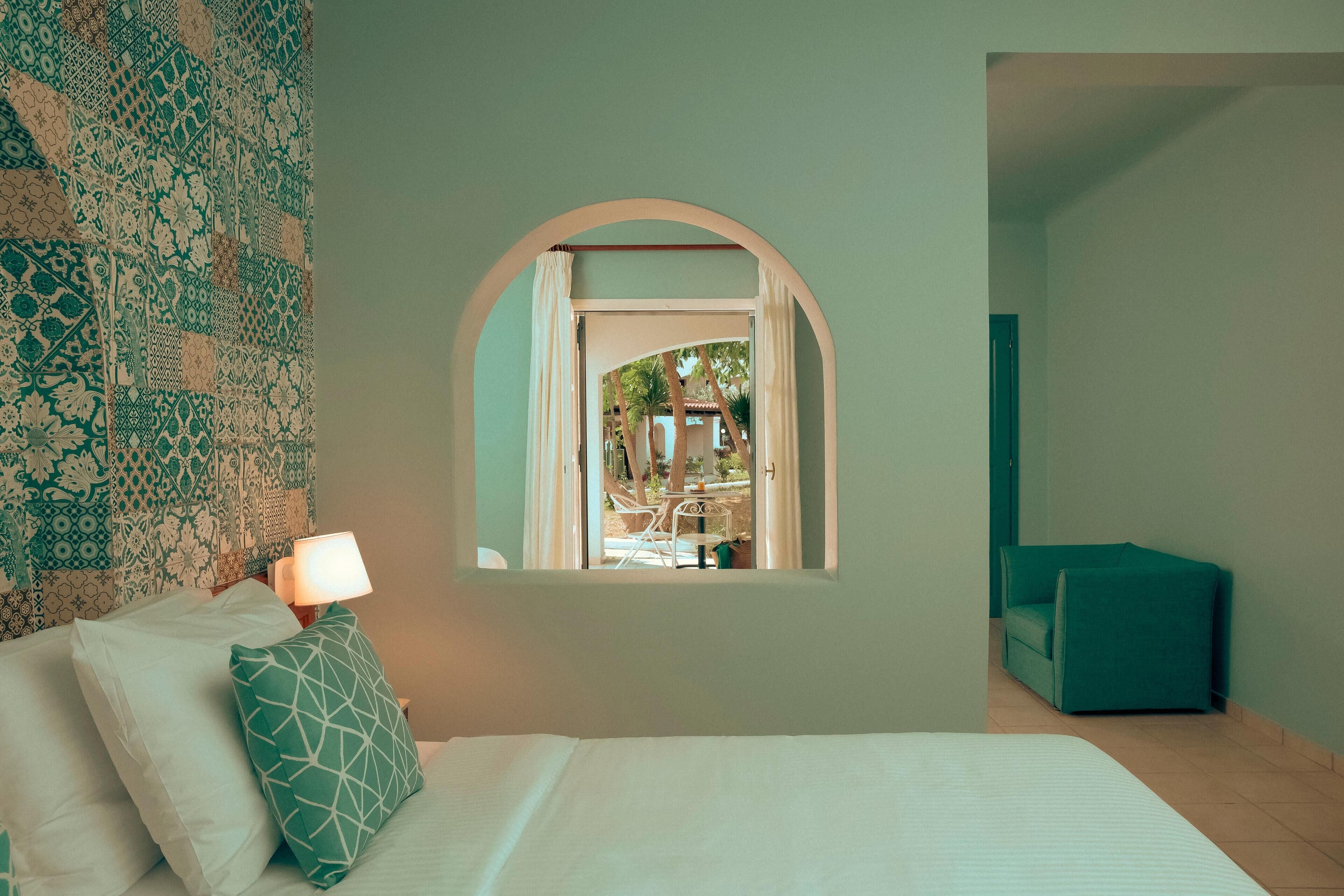 Un dormitorio con cama, una ventana en arco, una mesita de noche con lámpara y vistas a un espacio exterior con sillas y una mesa.