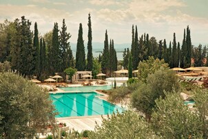 Una gran piscina rodeada de tumbonas y sombrillas, con altos cipreses y vistas a montañas distantes.
