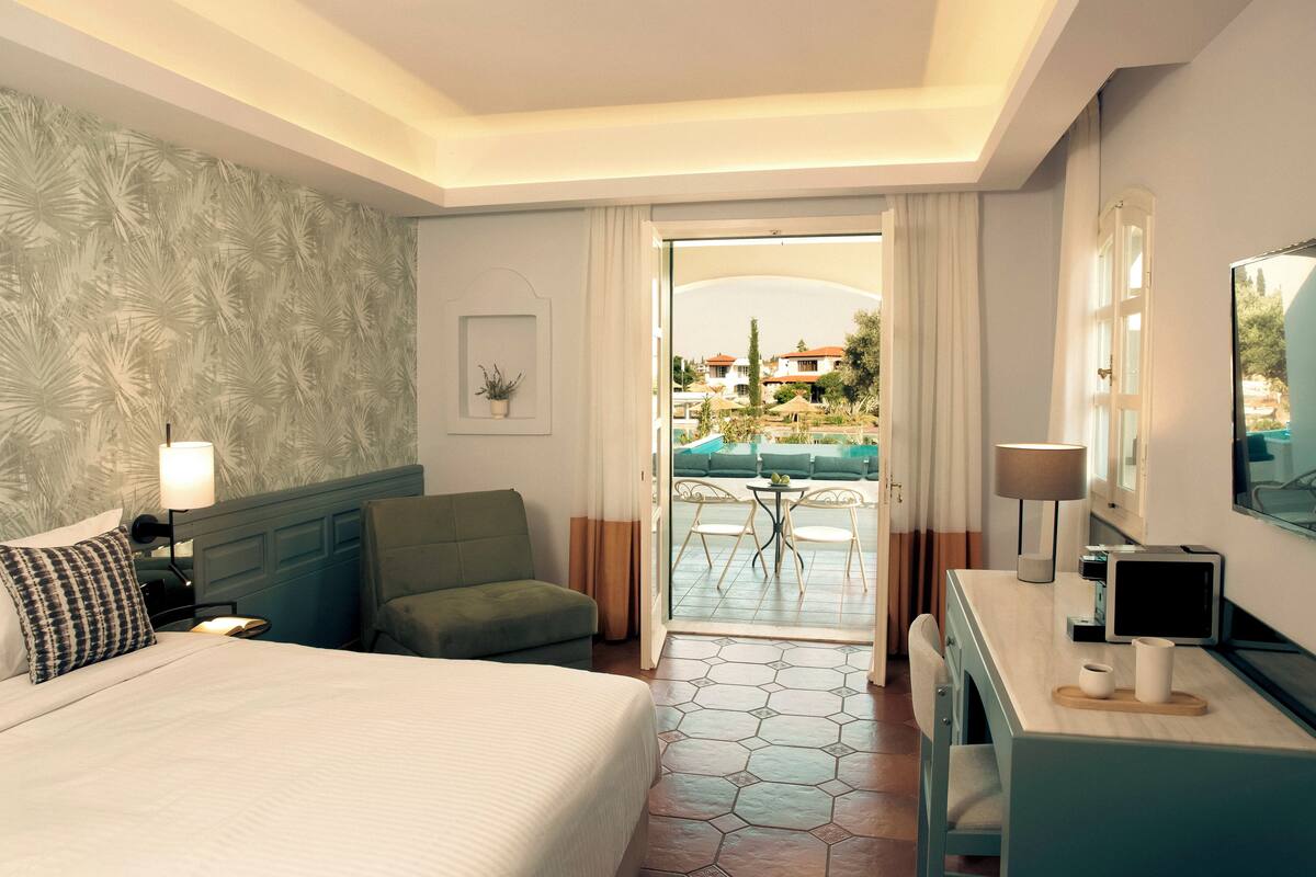 Een hotelkamer met een bed, een bureau, een stoel, een klein tafeltje en uitzicht op een buitenzwembad.
