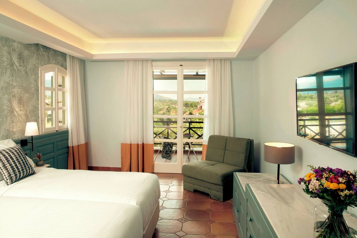 Een hotelkamer met een bed, een stoel, een bureau, een lamp, een vaas met bloemen en uitzicht op het omliggende landschap.