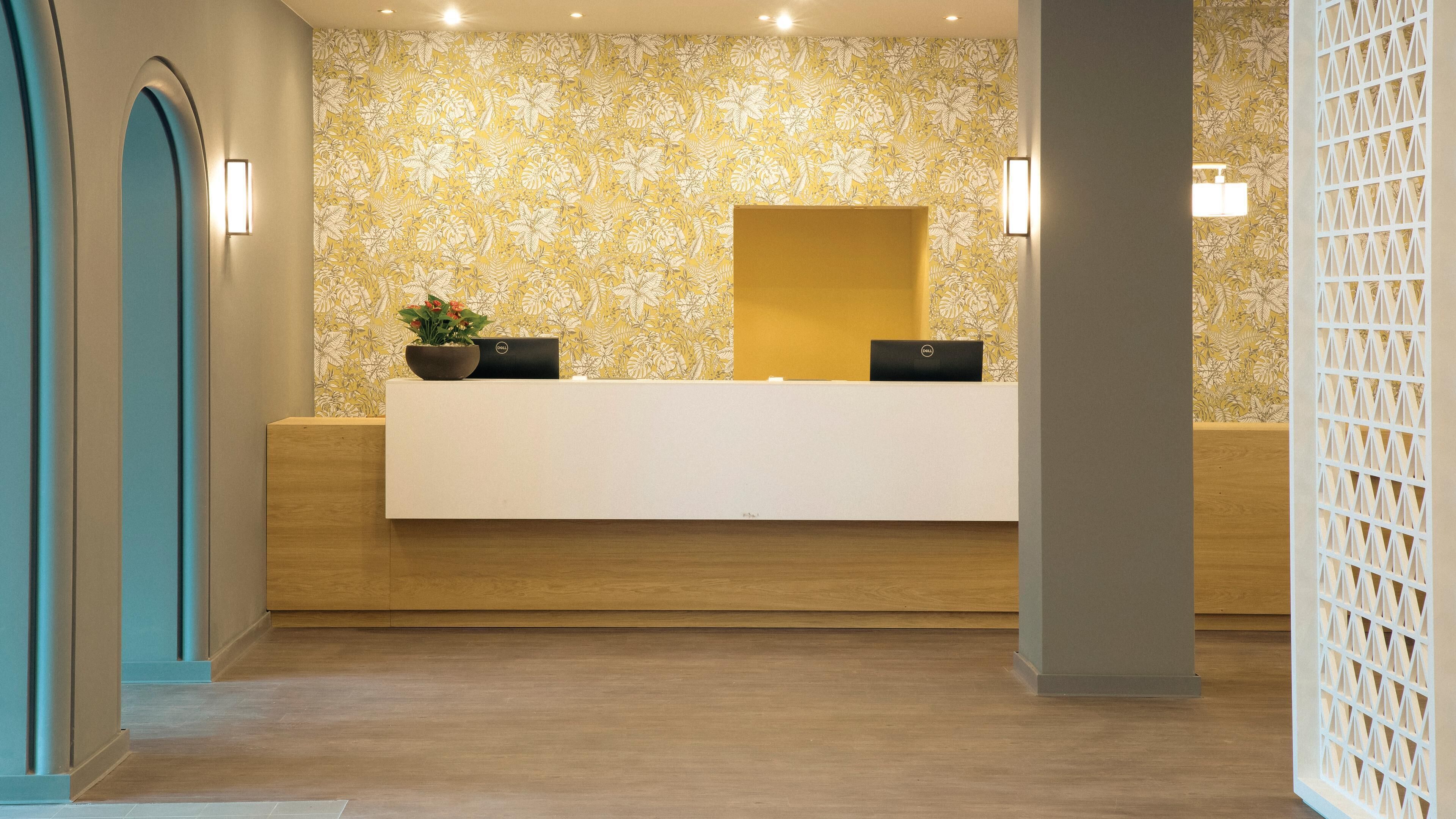 A reception desk with a floral wallpaper, a potted plant, and two laptops.