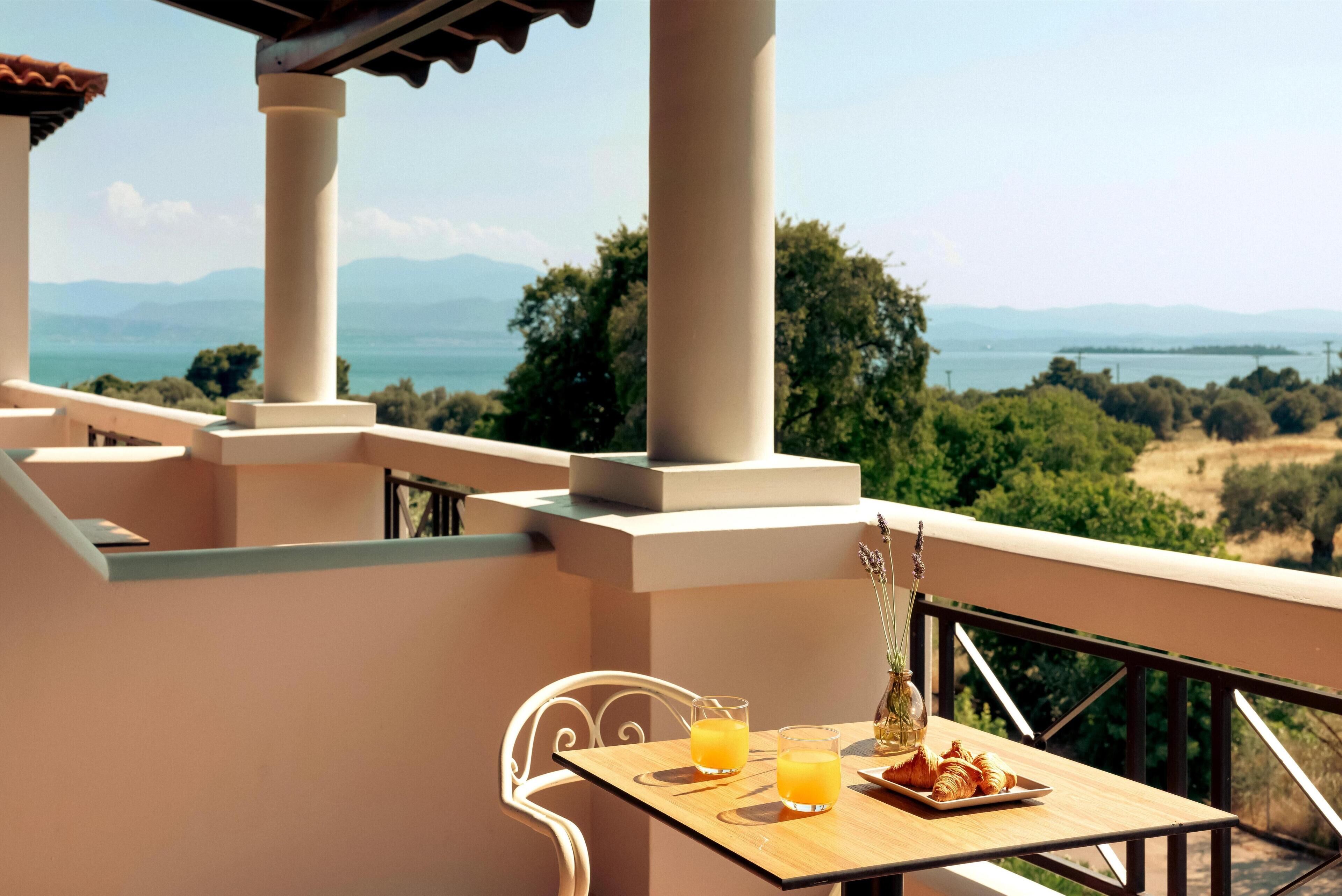 A balcony with a table set for two, two glasses of orange juice, a plate with croissants, and a vase with flowers.