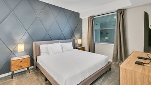Ein Hotelzimmer mit einem großen Bett, zwei Nachttischen mit Lampen, einem Holztisch, einem Fernseher und einem Fenster mit Vorhängen.