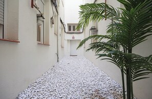 Une ruelle étroite pavée de petits cailloux blancs, une porte blanche au fond, et un pot de fleurs sur la droite.