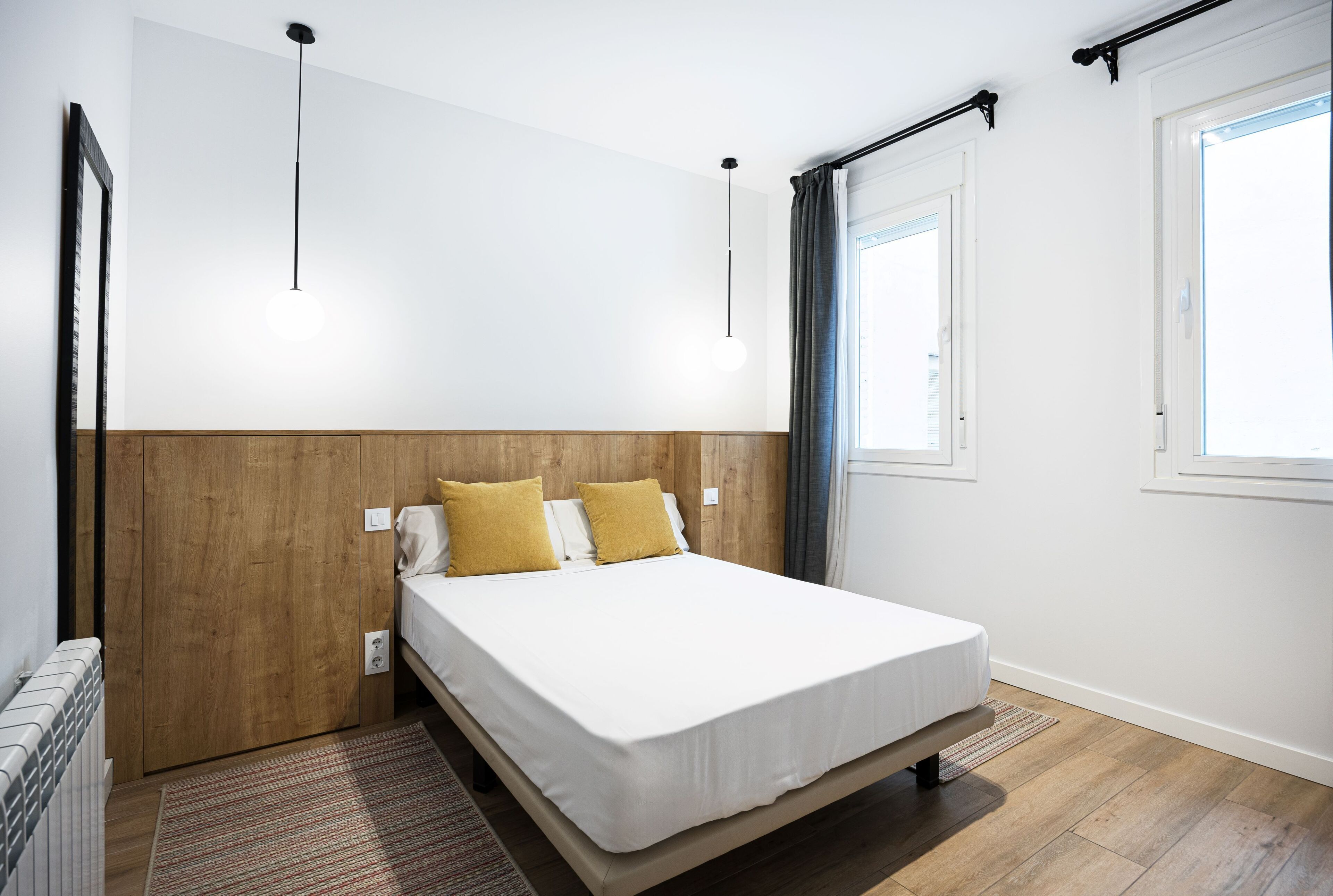 A bedroom with a wooden headboard, a white bedspread, and two pillows. There is a window with curtains, a radiator, and a hanging light fixture.