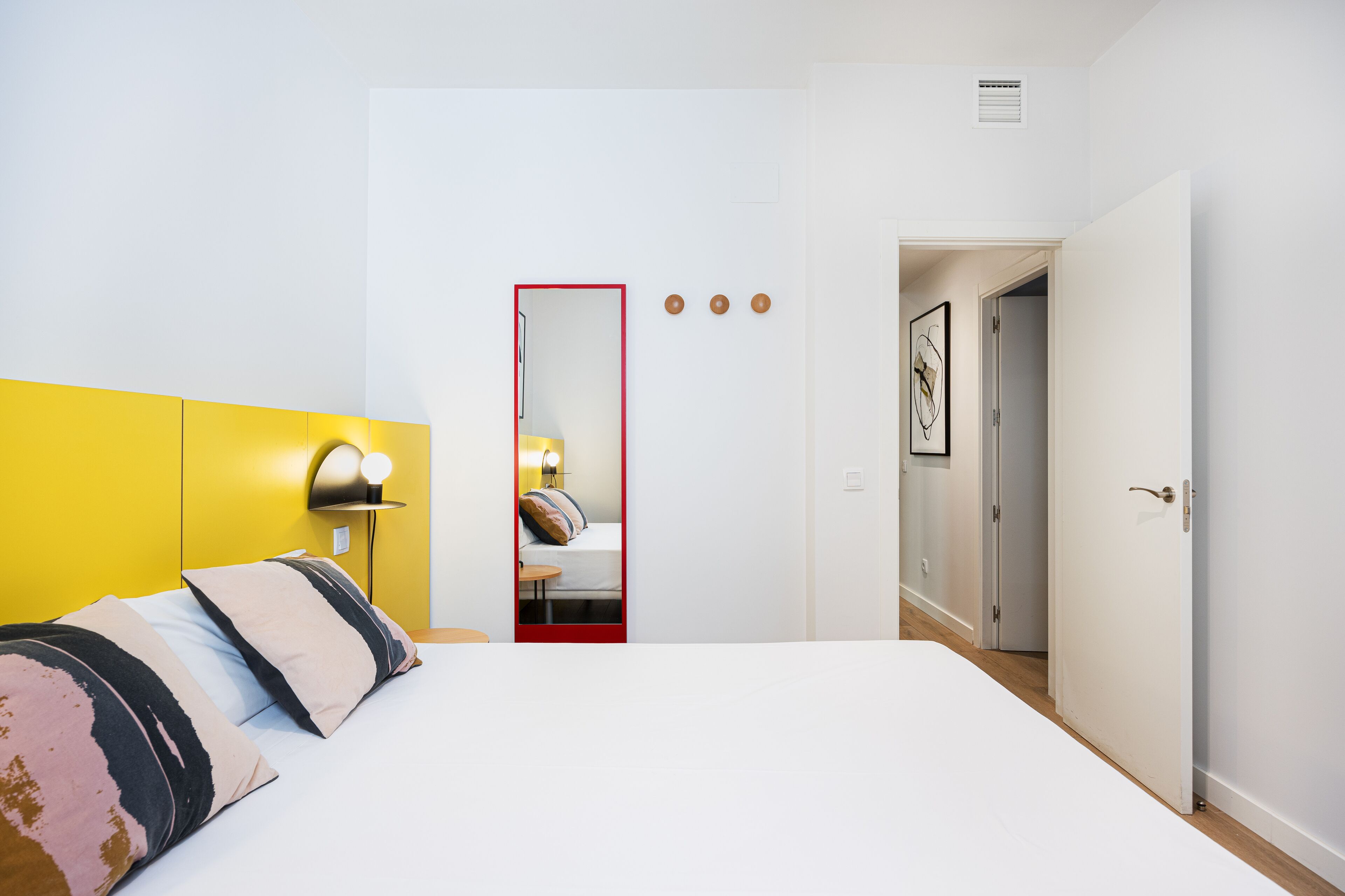 A hotel room with a bed, a mirror, a bedside lamp, and a door leading to another room.