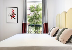 Une chambre à coucher avec un lit, une fenêtre avec des rideaux, une œuvre d’art abstraite encadrée, et une vue sur un balcon et la verdure à l’extérieur.