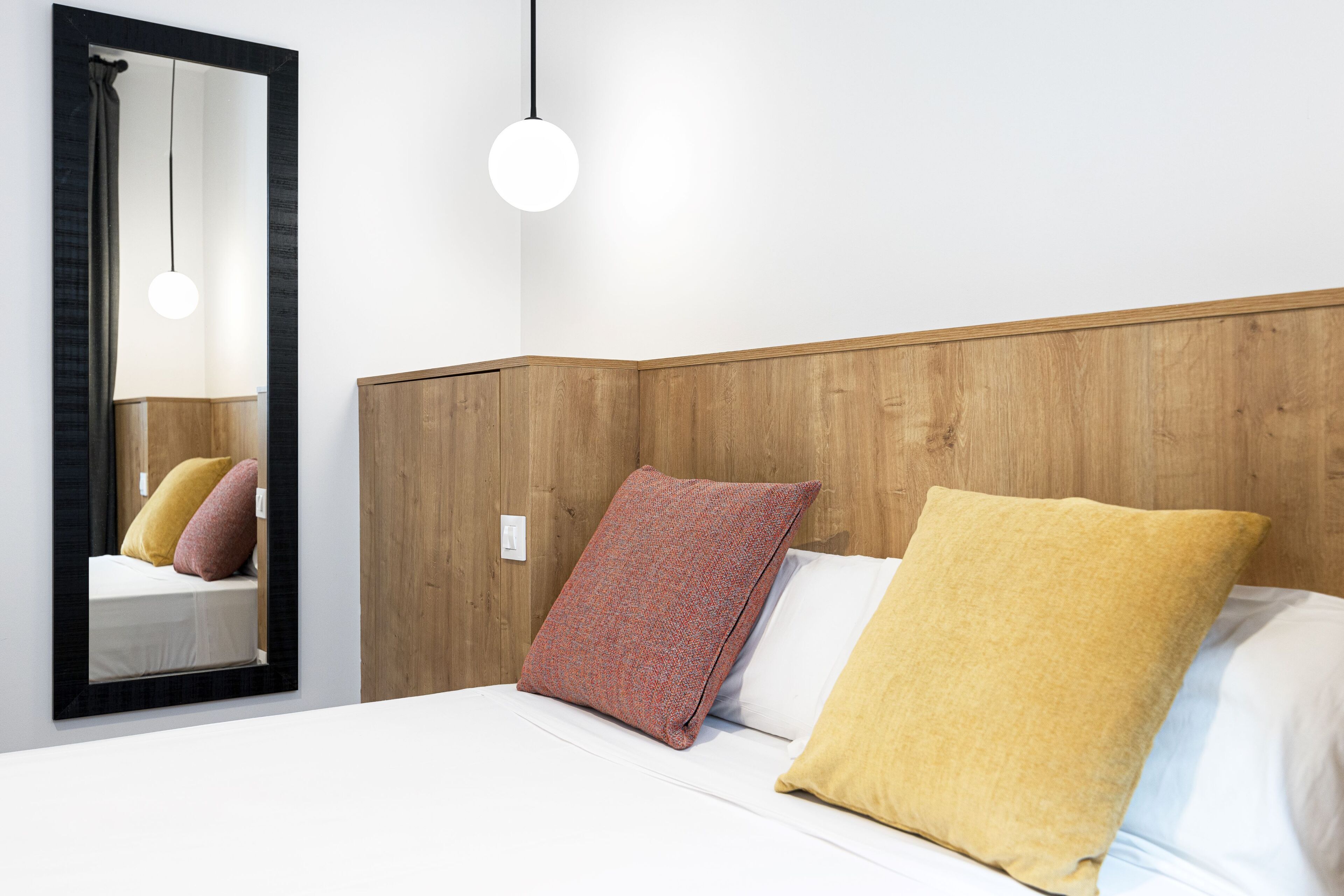 A neatly made bed with a wooden headboard, a mirror with a black frame, and a single hanging light fixture.