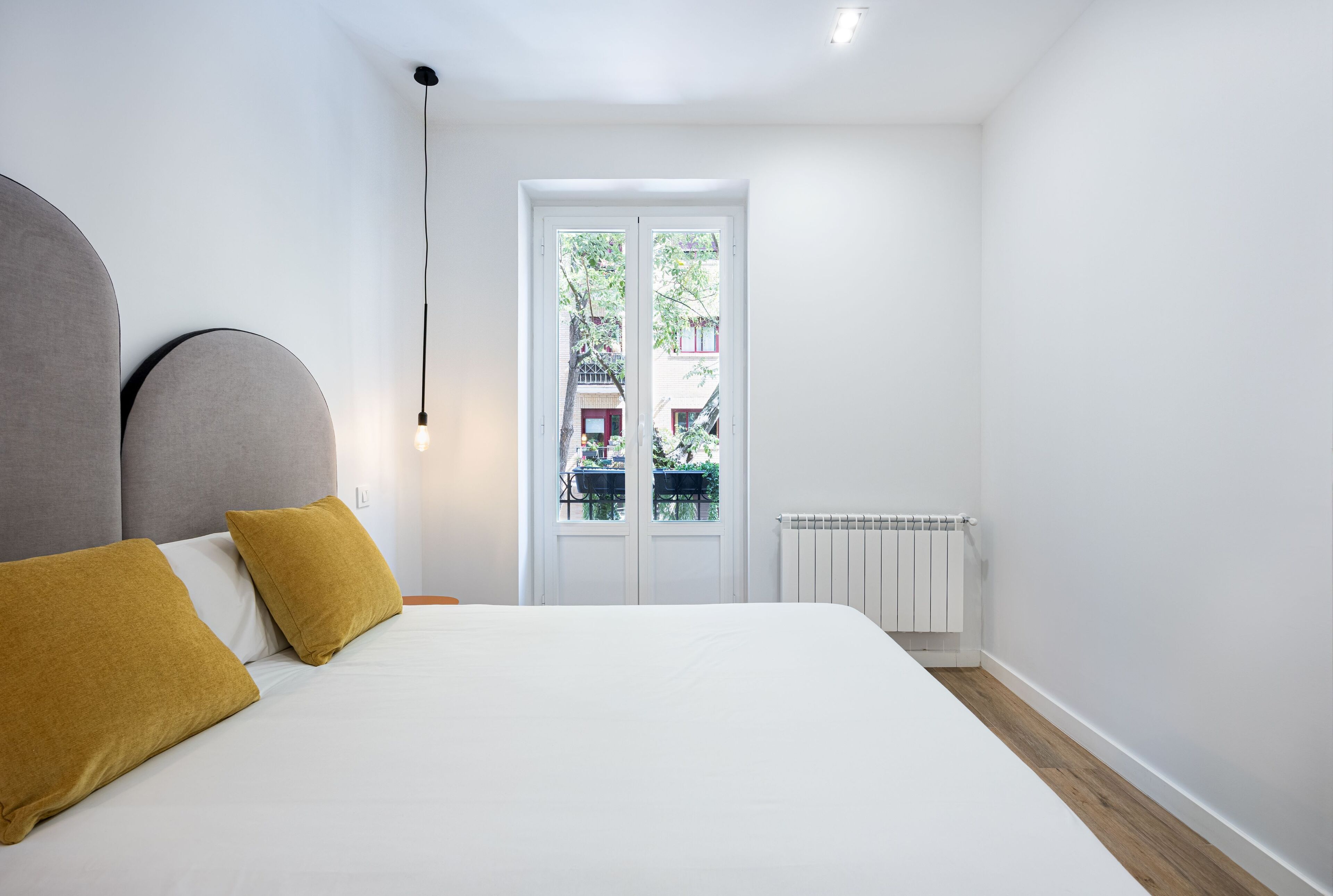 A bedroom with a bed, two mustard-colored pillows, a white headboard, a white radiator, and a window with a view of a balcony and buildings.