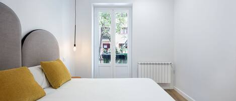 A bedroom with a bed, two mustard-colored pillows, a white headboard, a white radiator, and a window with a view of a balcony and buildings.