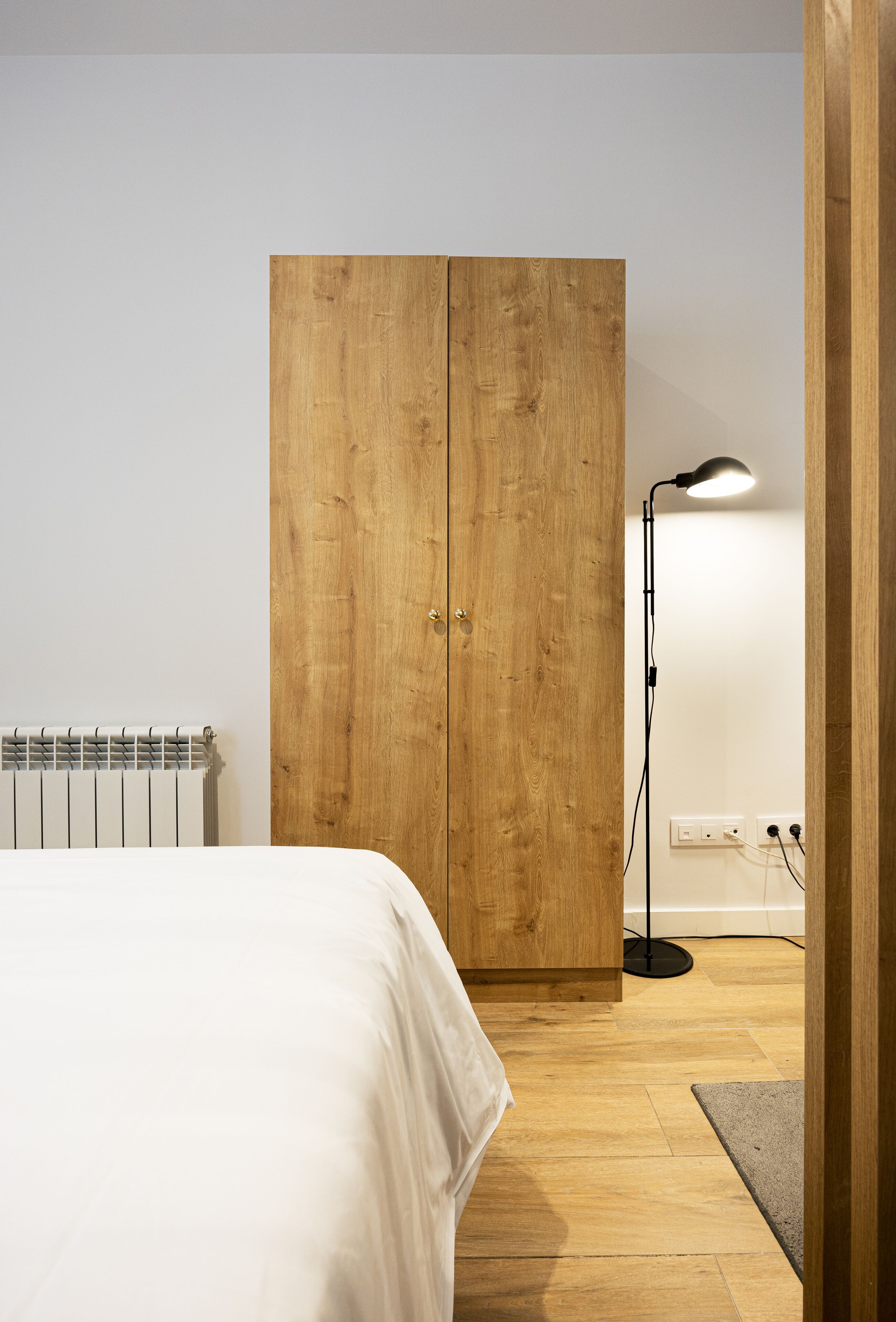Ein Schlafzimmer mit einem Holzkoffer, einem weißen Bett, einer Stehlampe und einer Heizung.