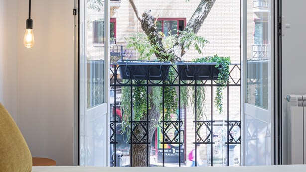 Un dormitorio con cama, una lámpara de noche y vistas a un balcón con plantas y una barandilla.