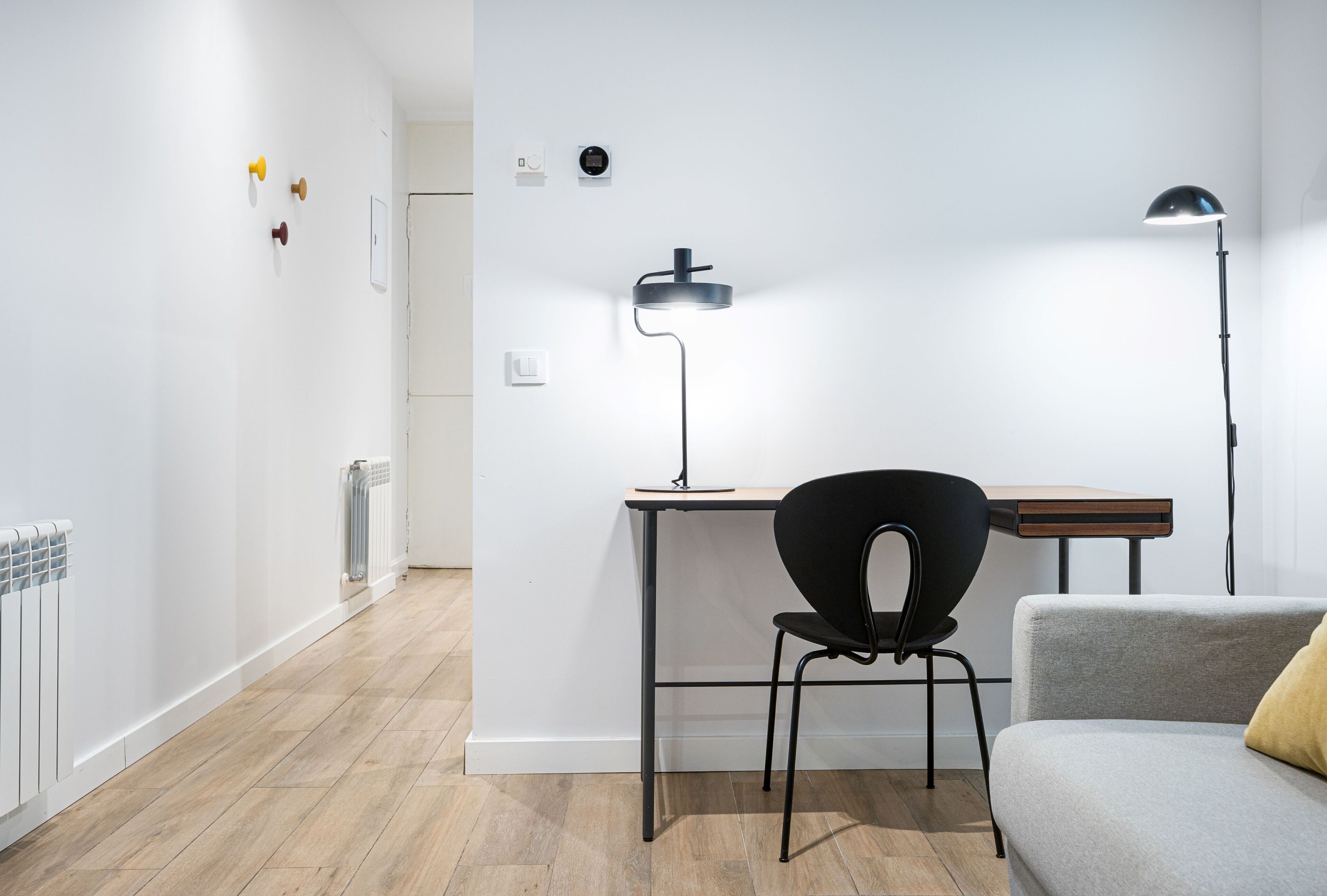 A modern, minimalist room with a wooden floor, a black chair, a grey sofa, and a desk with a lamp.