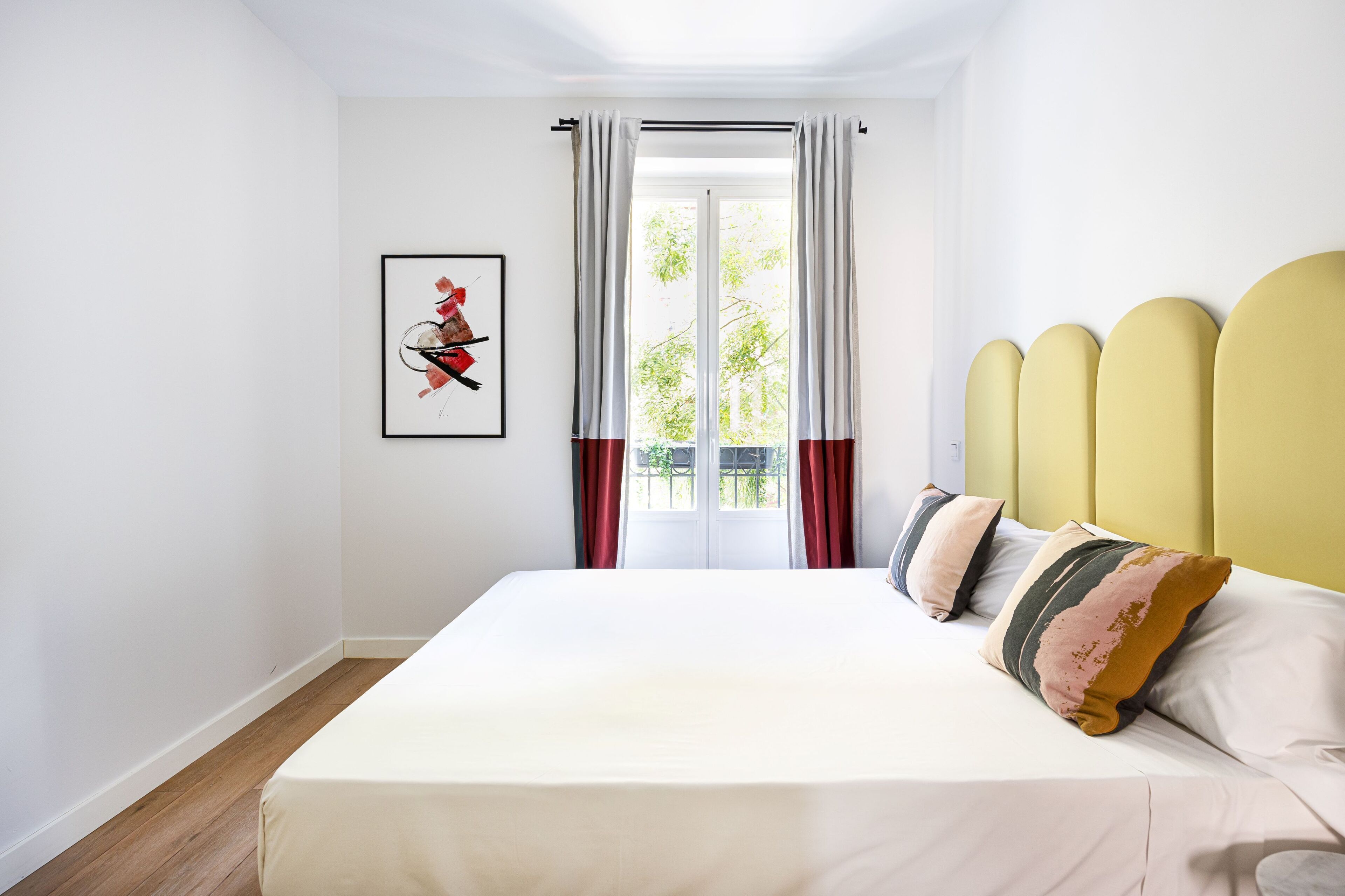 A bedroom with a bed, a window with curtains, a framed picture on the wall, and a yellow headboard.