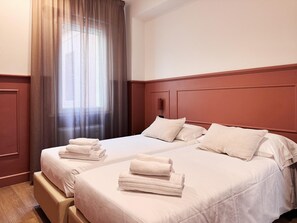 Habitación de hotel con dos camas, cabecero de madera, ventana con cortinas y una pila de toallas sobre cada cama.