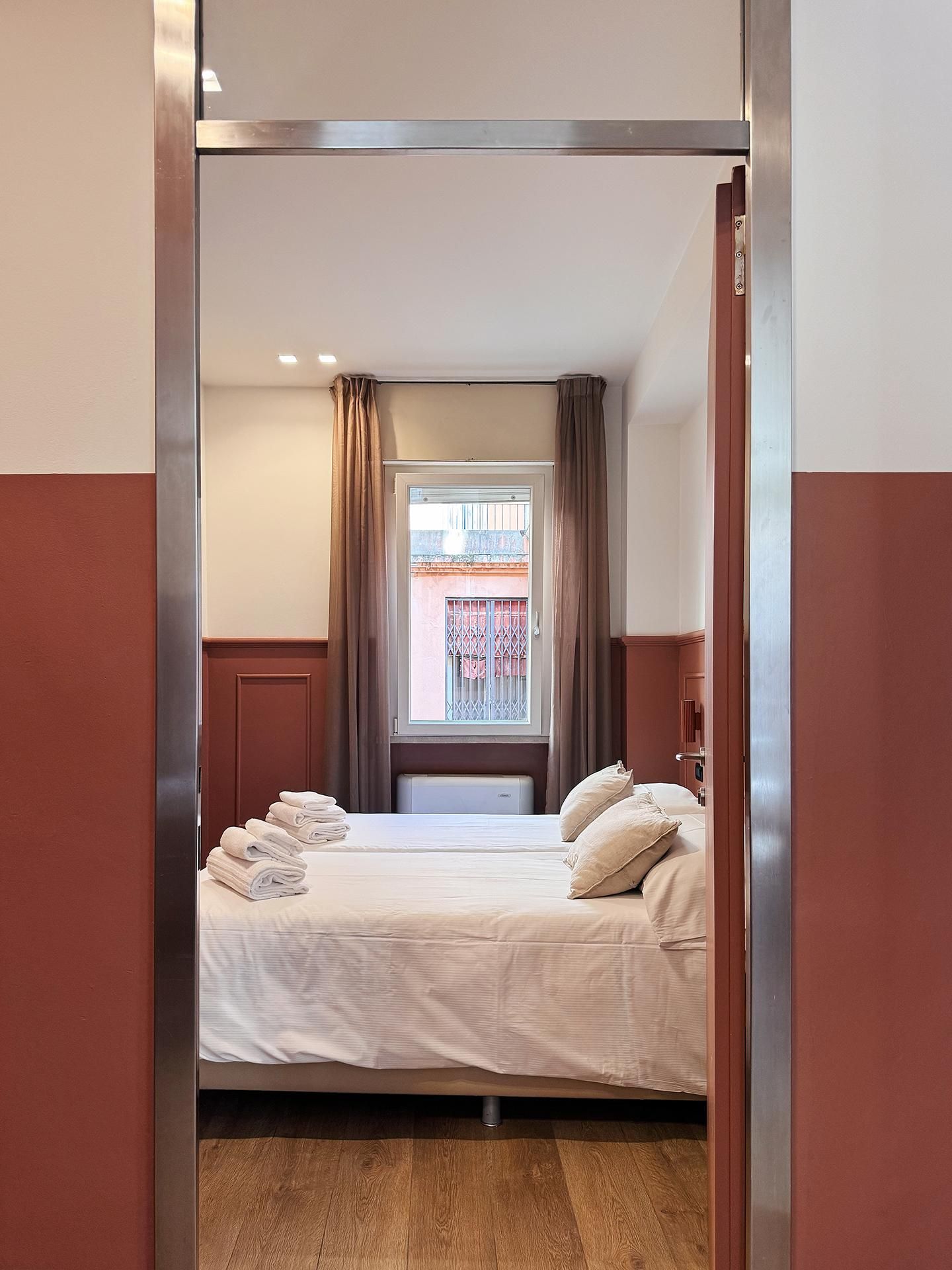A hotel room with a bed, a window with curtains, a view of the outside, and a wooden floor.