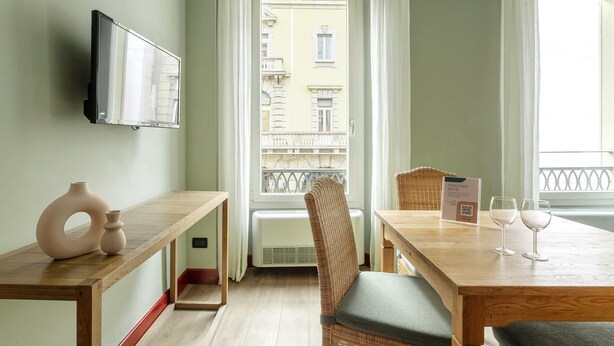 A hotel room with a wooden table, wicker chairs, a TV mounted on the wall, and a balcony with a view of buildings. - Mercanzia apartment. (Bologna)