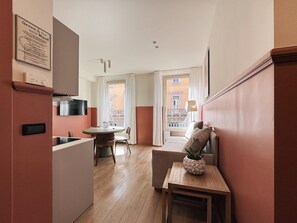 Un couloir étroit mène à un espace repas comprenant une table et des chaises, une petite table ornée d’un objet décoratif, et une vue sur un bâtiment à travers une porte ouverte.