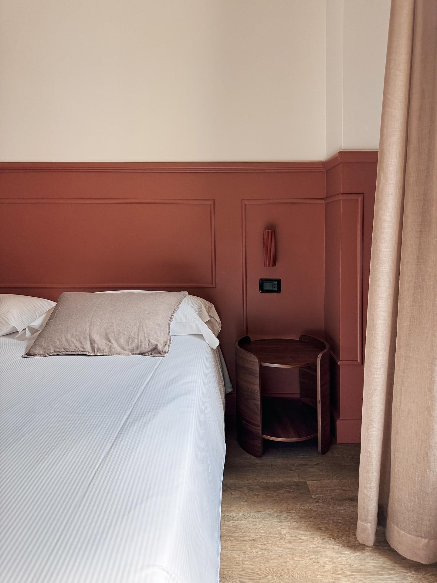 A hotel room with a bed, a small wooden side table, and a wall-mounted control panel.