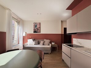 Une chambre d’hôtel de taille réduite, comprenant un lit, une table à manger, un coin cuisine et une fenêtre avec des rideaux.