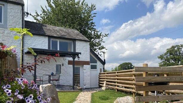 A stone house with a wooden fence, a stone pathway, and flowering plants. - Delightful One Bed Lake District Cottage (Penrith)