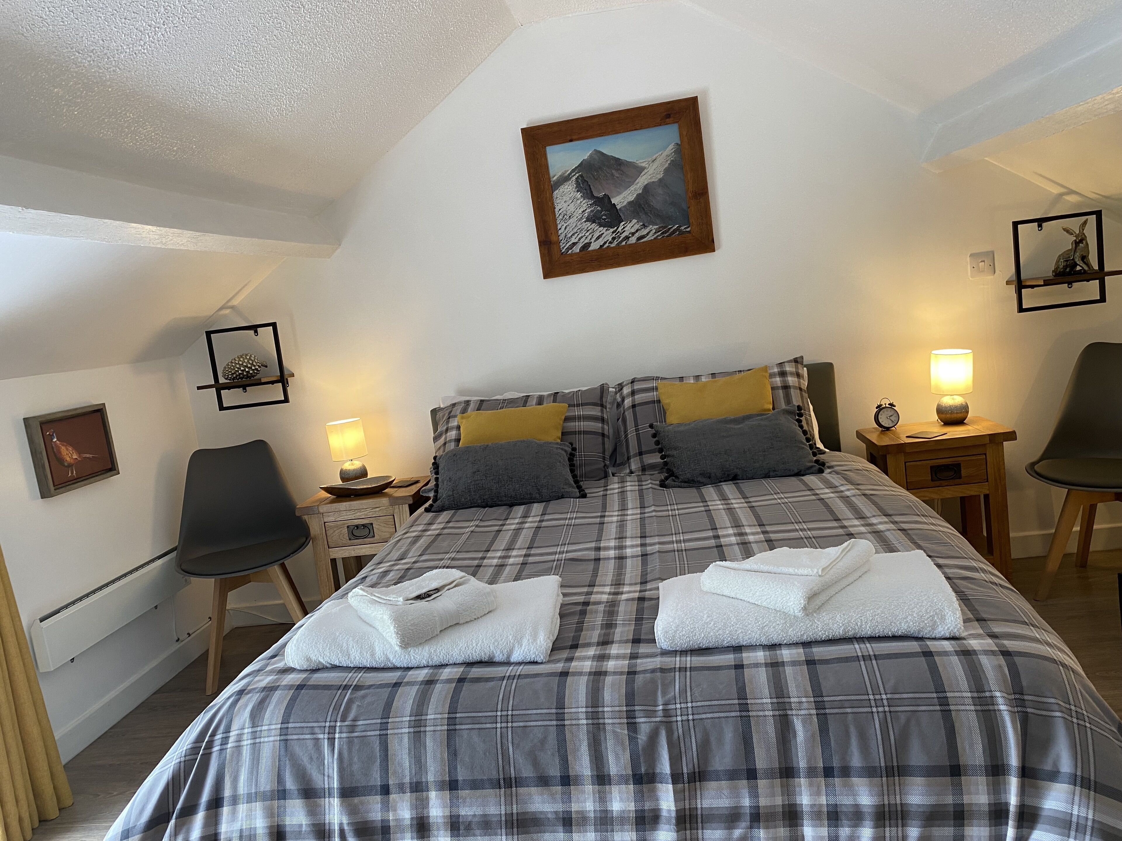 Ein ordentlich gemachtes Bett mit Karo-Bettwäsche, ein gerahmtes Bild darüber und ein kleiner Nachttisch mit Lampe und Uhr.
