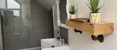 A modern bathroom with a glass shower enclosure, a white sink, and a wooden shelf holding two potted plants.