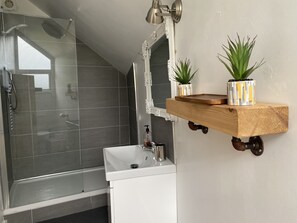 A modern bathroom with a glass shower enclosure, a white sink, and a wooden shelf holding two potted plants.