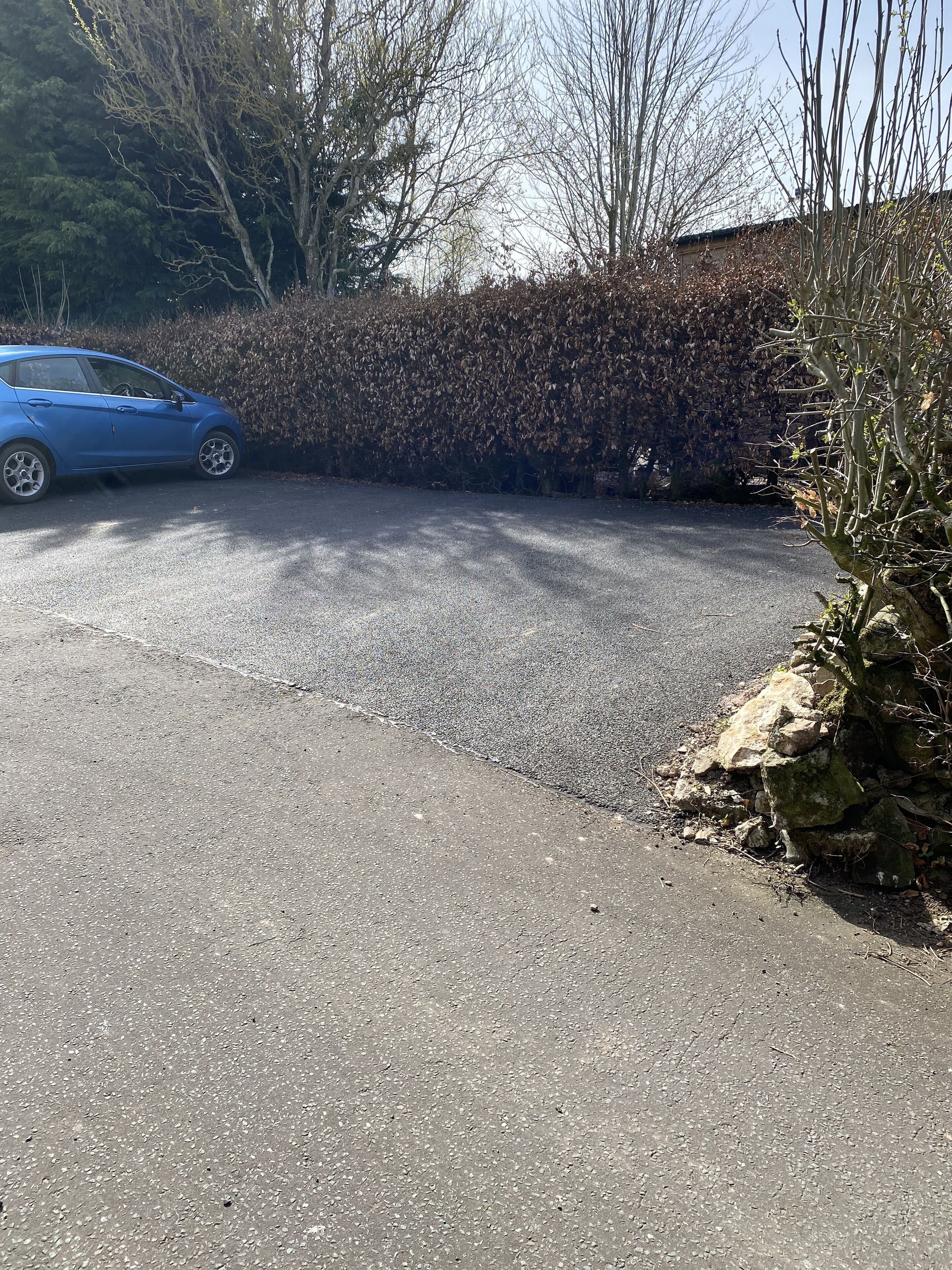 Un área de estacionamiento con un coche azul, un seto y una pared de piedra.