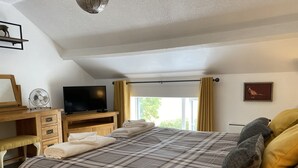 A bedroom with a bed, a desk with a television, a fan, and a window with curtains. - Delightful One Bed Lake District Cottage (Penrith)