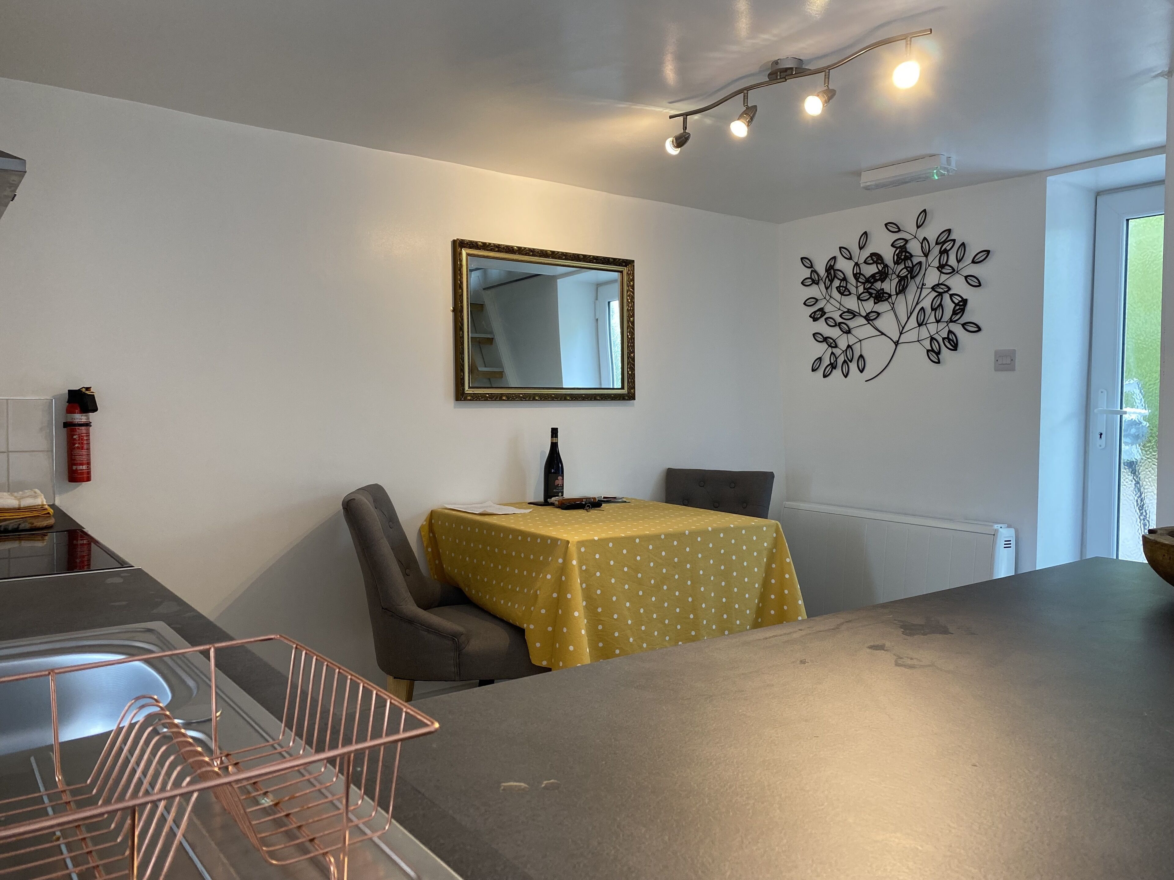 A dining area with a table covered in a yellow tablecloth, a chair, a mirror on the wall, and a decorative wall piece.