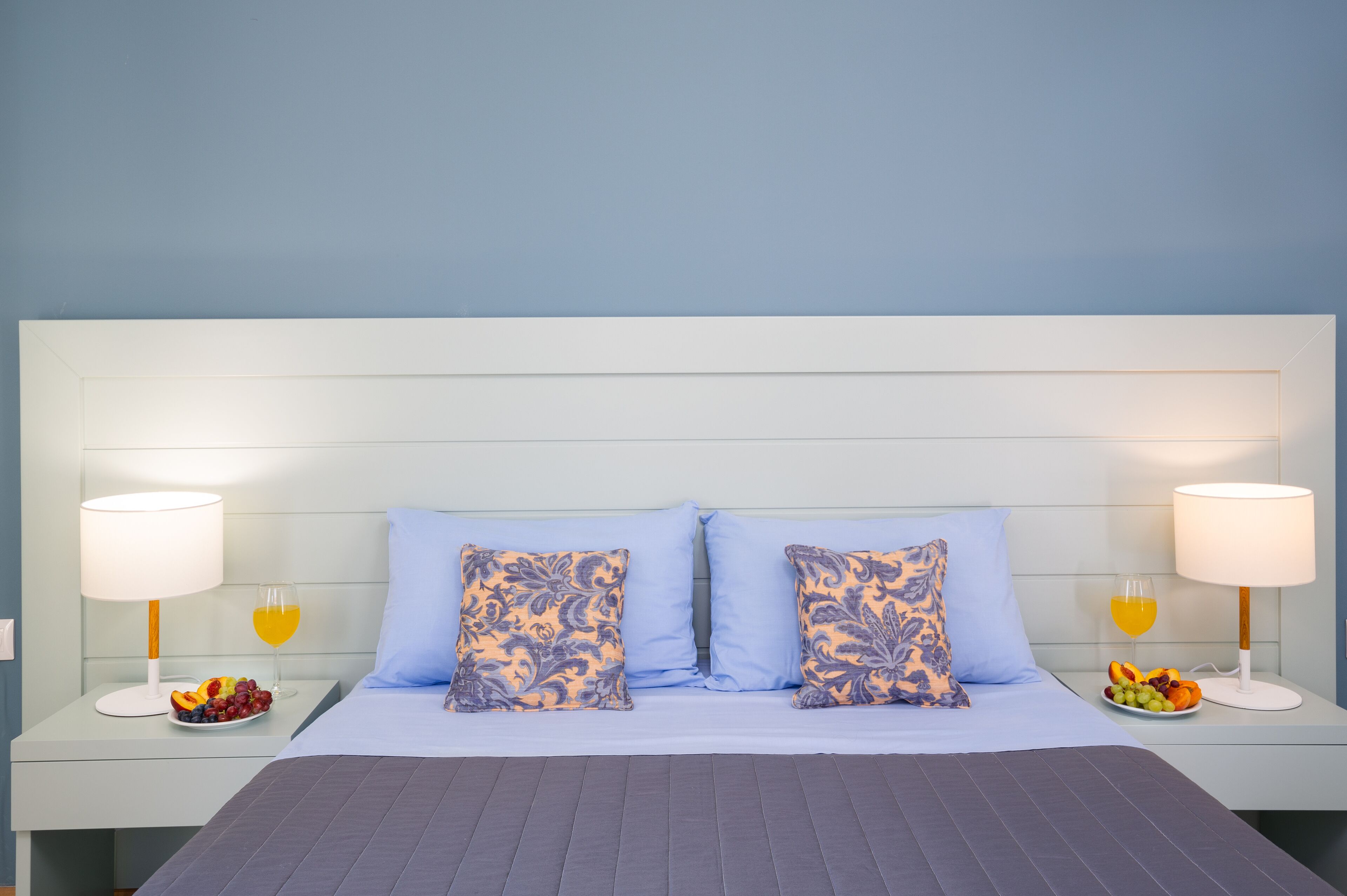 Una cama bien hecha con dos almohadas, una mesita de noche con frutero y un vaso de jugo de naranja, y una lámpara a cada lado de la cama.