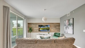 A modern living room with a large sofa, a flat-screen TV mounted on a wooden wall, and a contemporary pendant light. - 2bd/2ba condo close to Disney, Gated community at Storey Lake (Kissimmee)
