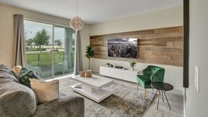 A modern living room with a large window, a flat-screen TV mounted on a wooden wall, a white console table, a green armchair, and a small side table. - 2bd/2ba condo close to Disney, Gated community at Storey Lake (Kissimmee)