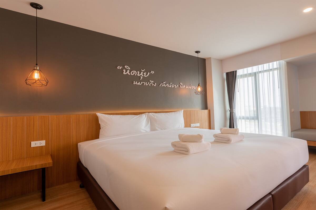 A modern hotel room with a large bed, a bedside table, a bench, and a wall with a light fixture and text.