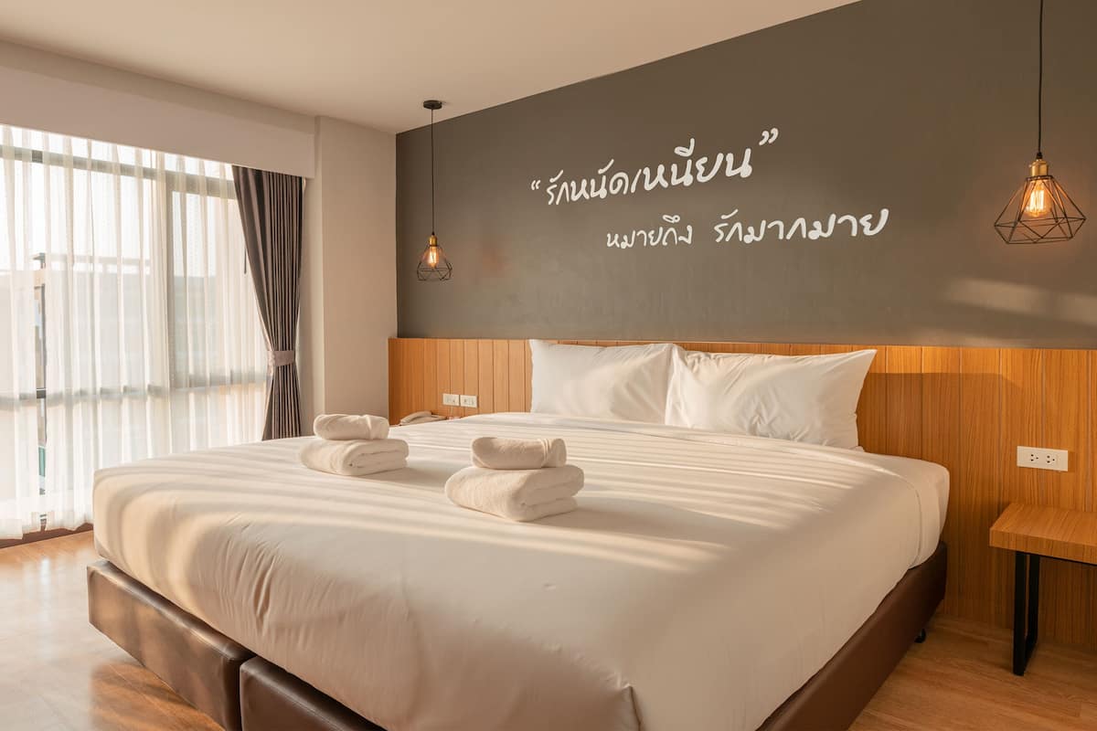 A modern hotel room with a large bed, wooden headboard, and a wall with Thai text.