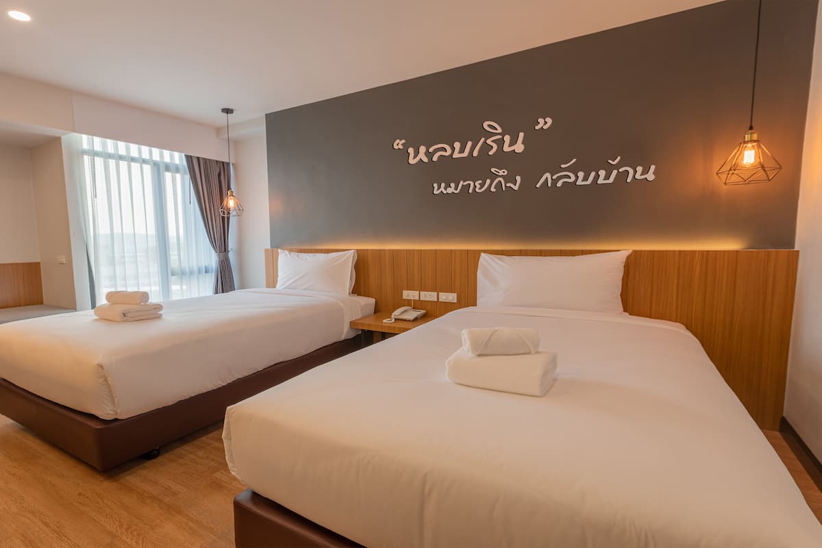 A hotel room with two beds, a bench, an air conditioning unit, and a wall with Thai text.