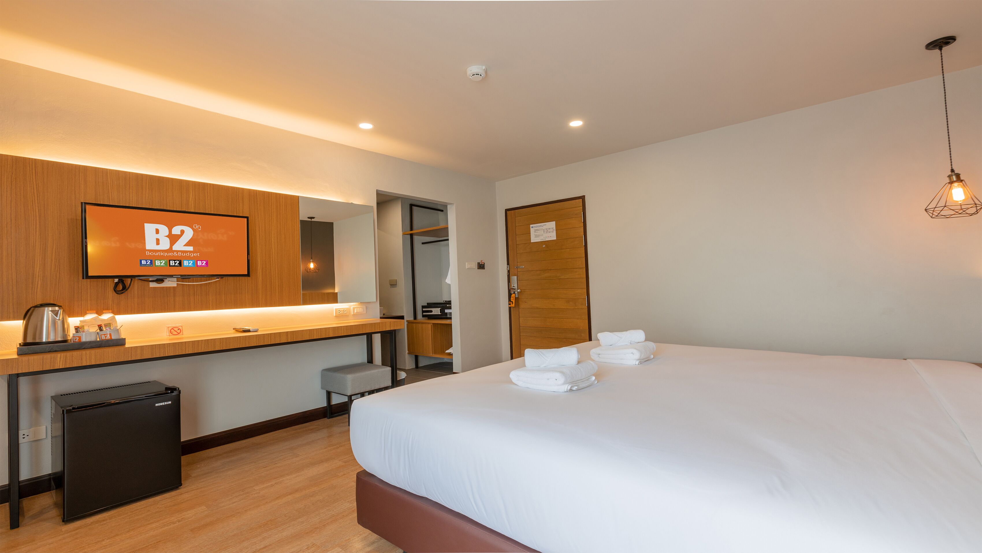 A hotel room with a bed, a desk with a computer, a television displaying 'B2 Boutique & Budget', a mirror, and a wooden door.