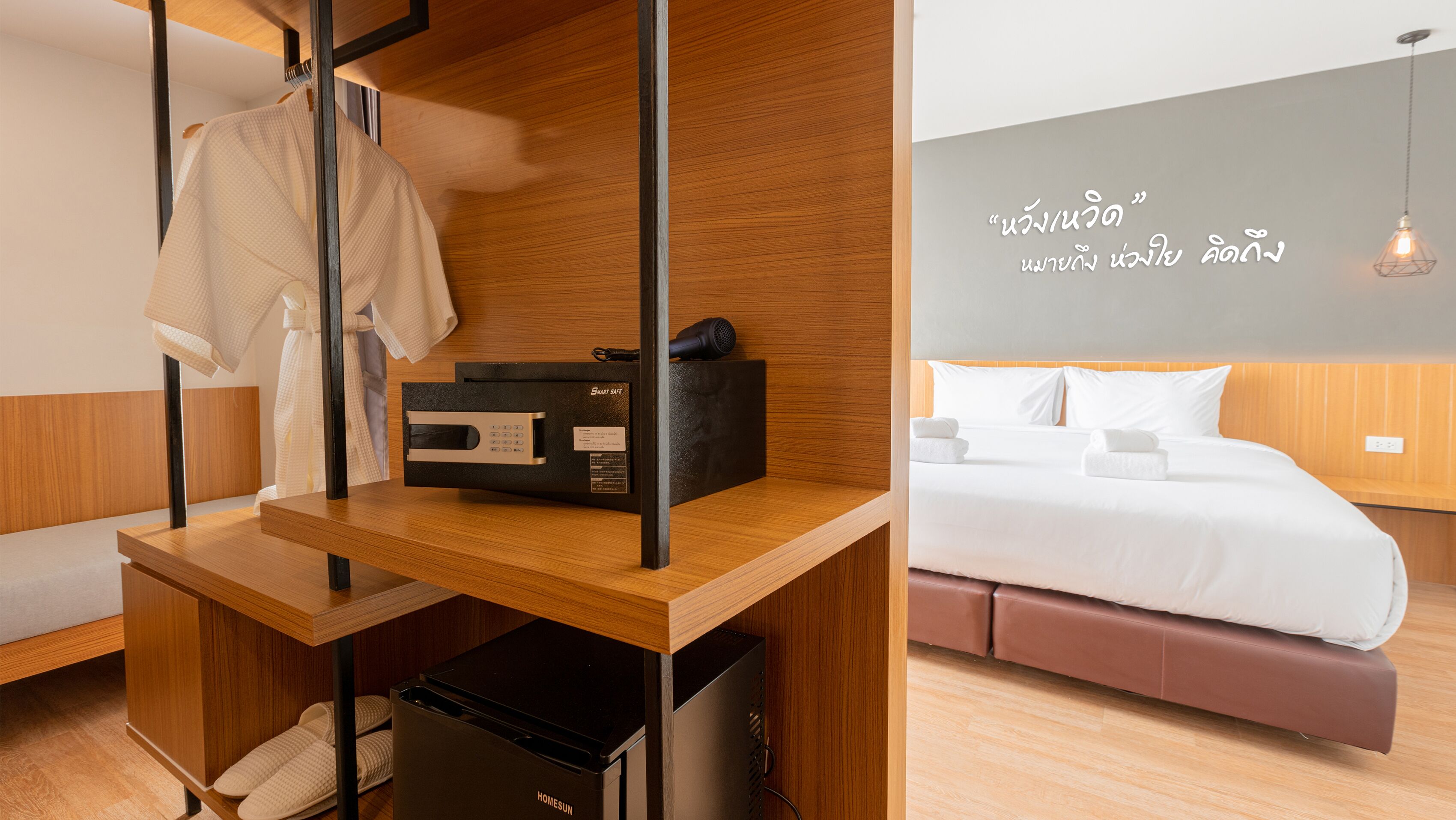 A hotel room with a wooden desk, a bed with white linens, and a wall with Thai text.