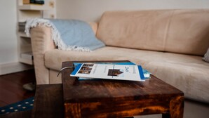 Una mesa de centro de madera con un folleto frente a un sofá color beige con un plaid azul.