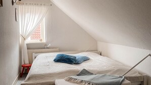 Un dormitorio con techo inclinado, una cama individual con cajones para guardar objetos, un taburete pequeño y una ventana con cortinas translúcidas.