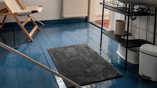 Un dormitorio pequeño y acogedor en el ático, con techo inclinado, una ventana con cortinas traslúcidas, un radiador, una silla de madera, un tapete blanco y negro, un lavamanos y un inodoro.