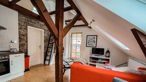 A cozy loft apartment with exposed wooden beams, a stone wall, a kitchen with white cabinets, a red sofa, and a TV on a white stand.