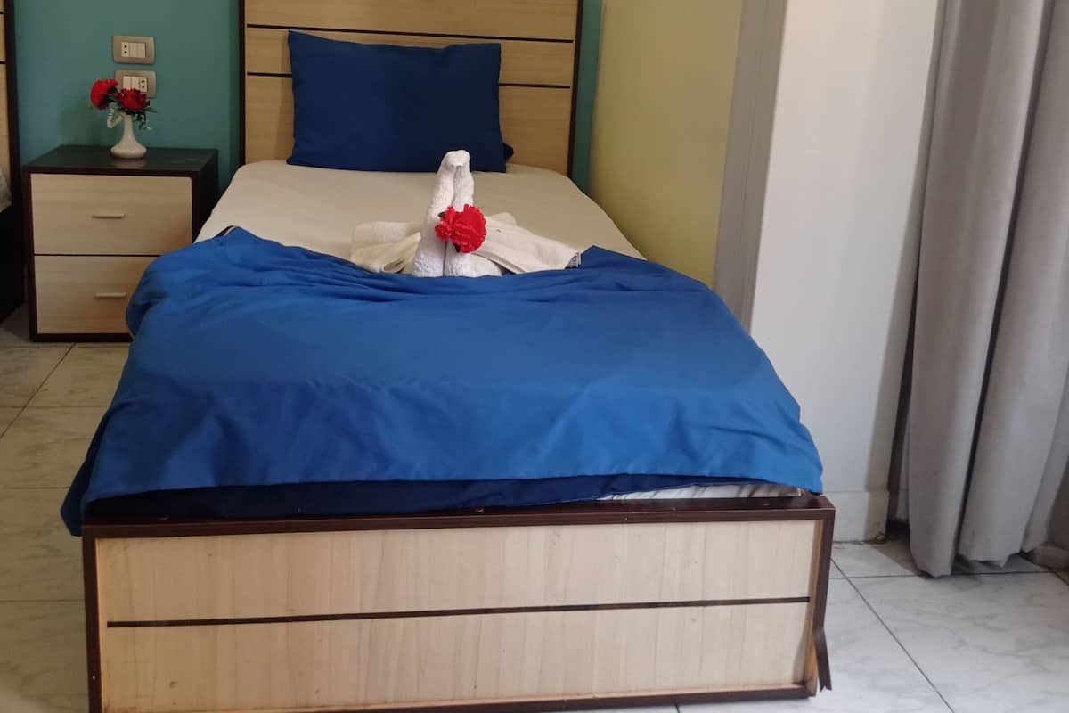 A neatly made bed with blue bedding and a white pillow, a nightstand with a vase of red flowers, and a wall-mounted light fixture.