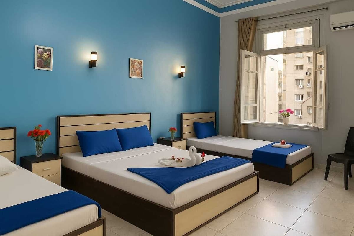 A hotel room with two beds, a window with a view of buildings, and wall-mounted lights.